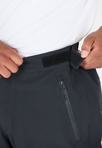 Pantalons noirs avec une taille en Velcro et une poche zippée sur le côté droit, fabriqués à partir d'un tissu lisse et extensible.