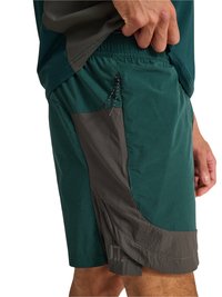 Mand justerer elastisk linning på mørkegrønne og grå træningsshorts med lynlåslomme i siden.