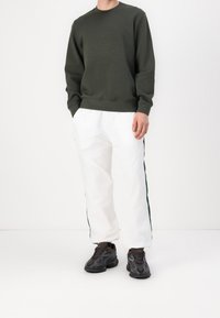 Sweatshirt vert foncé à col rond avec un logo texturé, associé à un pantalon blanc avec des bandes latérales vertes et des baskets noires à motif.