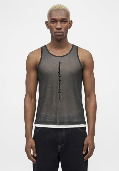 Μαύρο αμάνικο τοπ από δίχτυ με κάθετο μικρό κείμενο που φοριέται από άτομο με κοντά ξανθά μαλλιά και σκουρόχρωμο παντελόνι σε λευκό φόντο.