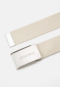 Cinta de tecido bege com fivela de metal prateado gravada com "Dickies", disposta em plano sobre fundo branco.