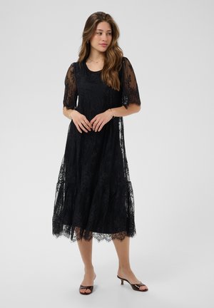 Femme aux longs cheveux ondulés portant une robe midi noire en dentelle avec des manches courtes transparentes et des escarpins noirs à bout ouvert, debout devant un fond uni.