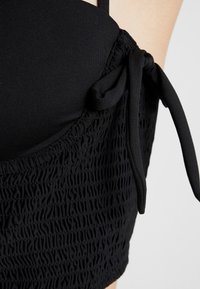 Černé plavky s hladkým materiálem na vršku a texturovaným, nabíraným materiálem dole. Obsahují nastavitelná ramínka a detailem v podobě uzlu na boku.