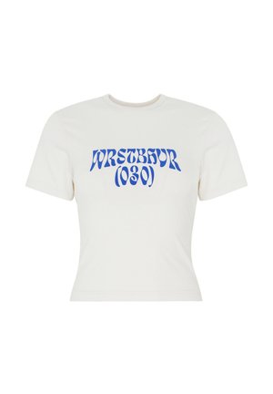 Femme portant des lunettes de soleil noires, un t-shirt court blanc avec un texte bleu, un jean large et sombre avec ceinture, tenant une veste noire dans une main.