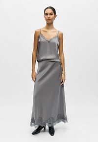 Robe nuisette en satin gris avec bordure en dentelle, dotée de fines bretelles et d'une silhouette maxi fluide, associée à des bottines à talons noires texturées.