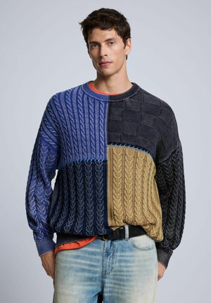 Joven con un suéter de punto grueso combinado en azul, negro y marrón claro, con vaqueros de lavado claro y una camisa naranja debajo.
