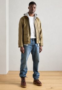 Ελαιόχρωμο αδιάβροχο μπουφάν με γκρίζα φόδρα κουκούλας, μπροστινές τσέπες, συνδυασμένο με ανοιχτό μπλε τζιν και καφέ δερμάτινα μποτάκια σε ξύλινο πάτωμα.