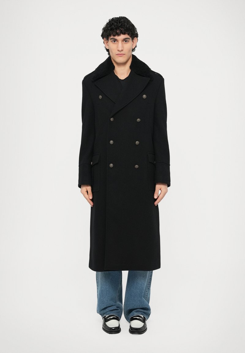 Abrigo negro de lana de doble botonadura con un amplio cuello tipo chal, grandes botones y bolsillos laterales. Llevado sobre unos vaqueros azules de pierna ancha y zapatos negros.