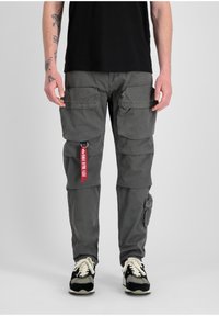 Los pantalones cargo de tela gris cuentan con múltiples bolsillos frontales, un bolsillo lateral y una etiqueta roja. Están combinados con zapatillas en blanco y negro.
