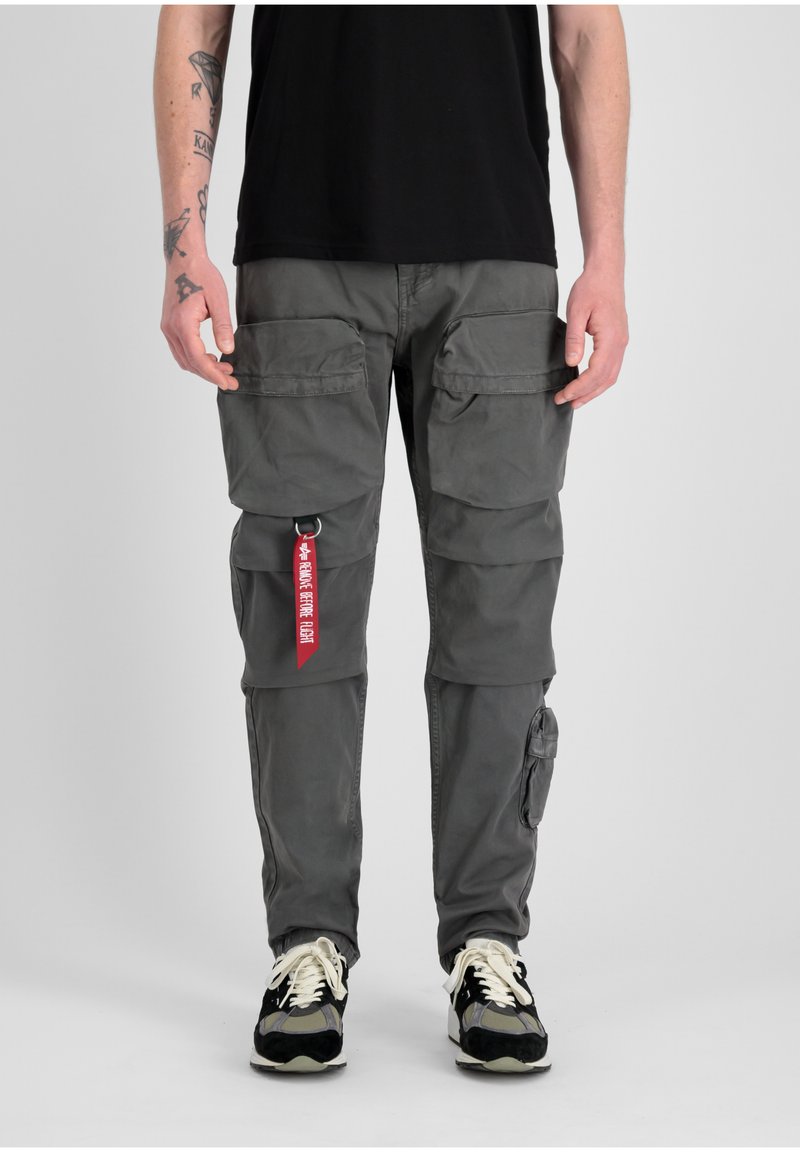 Los pantalones cargo de tela gris cuentan con múltiples bolsillos frontales, un bolsillo lateral y una etiqueta roja. Están combinados con zapatillas en blanco y negro.