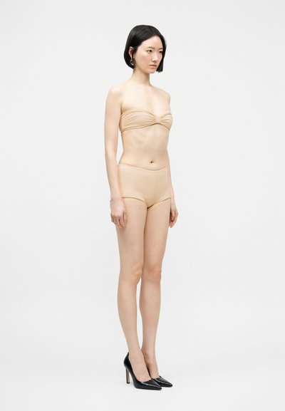 Femme aux cheveux noirs courts portant un haut bandeau beige, un short assorti et des talons hauts noirs, debout devant un fond clair uni.