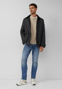 Mann trägt eine schwarze Jacke über einem beigen Pullover, blaue Jeans und weiße Turnschuhe, steht vor einem schlichten hellgrauen Hintergrund.