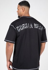 Fekete póló, amelyen a "GORILLA WEAR" felirat látható álcázó mintával, rövid ujjal, kontrasztos varrással és laza fazonnal.