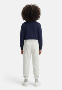 Άτομο με σγουρά μαλλιά στέκεται γυρισμένο προς τα πίσω, φοράει μπλε σκούρο φούτερ, ανοιχτό γκρι παντελόνι φόρμας με μικρό κείμενο στον αριστερό μηρό, γκρι κάλτσες και άσπρα αθλητικά παπούτσια.