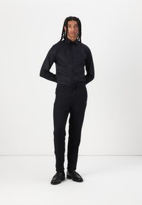Black button-up shirt with long sleeves, paired with tailored dark trousers and black dress shoes, set against a white background.