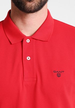 Hombre con una camisa polo roja de GANT con dos botones blancos y un pequeño logo bordado en el pecho izquierdo sobre un fondo blanco.