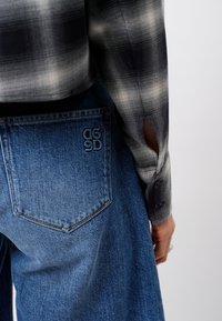 Plaid shirt with gray and white tones, unbuttoned cuff, paired with medium-wash blue jeans featuring a visible pocket logo.