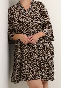 Vestido de estampado de leopardo con escote en V, mangas de tres cuartos, dobladillo con volantes y tejido ligero. Presenta manchas negras y marrones sobre un fondo tostado.