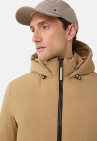 Beige Kapuzenjacke mit vollem Reißverschluss, hohem Kragen und Toggle-Akzenten. Kombiniert mit einer hellbraunen Mütze aus strukturiertem Stoff.