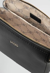Sac en cuir noir avec une surface texturée, fermeture éclair dorée, et un intérieur beige à motifs avec des accents de logo. "LIUJO" estampé sur l'extérieur.