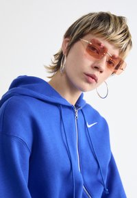 Zilā zip-up kapuce ar balto Nike logo, apvienota ar pārāk lielām rozā toni taisnstūra saulessargām un sudraba gredzenveida auskariem.