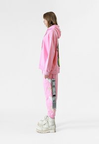 Sudadera rosa con un gráfico colorido en la espalda, combinada con joggers rosas a juego que tienen acentos texturizados y cordones blancos. Botas blancas.