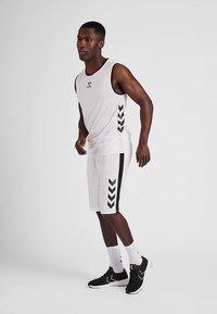 Vit linne med svart logotyp och fiskbensmönster på sidan, kombinerad med vita shorts med svarta detaljer, bärs med svarta sneakers.