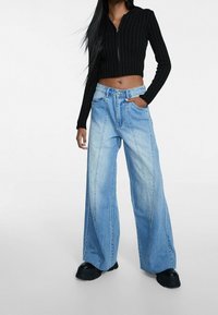 Svart ribbad kort zip-up sweatshirt och ljusblå vida jeans med framfickor och sömnadsdetaljer, kombinerat med svarta chunky skor.