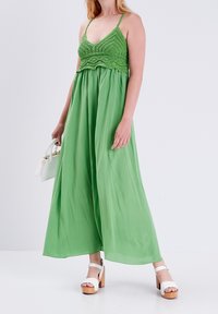 Femme portant un haut crochet vert et une jupe fluide avec des sandales compensées blanches, tenant un petit sac à main blanc devant un fond uni.