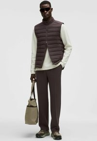 Gilet imbottito marrone sopra un maglione chiaro, abbinato a pantaloni scuri e sneakers con dettagli verdi. Accessorio: borsa a tracolla beige.