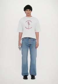 White oversized t-shirt with red graphic print, paired with blue flared jeans. Black shoes and holding a metallic accessory.