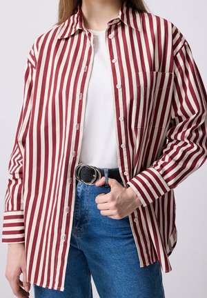 Osoba ubrana w koszulę zapinaną na guziki z pionowymi czerwono-białymi paskami, pod którą ma białą koszulkę, niebieskie dżinsy z czarnym paskiem z podwójną klamrą, ręka w kieszeni.