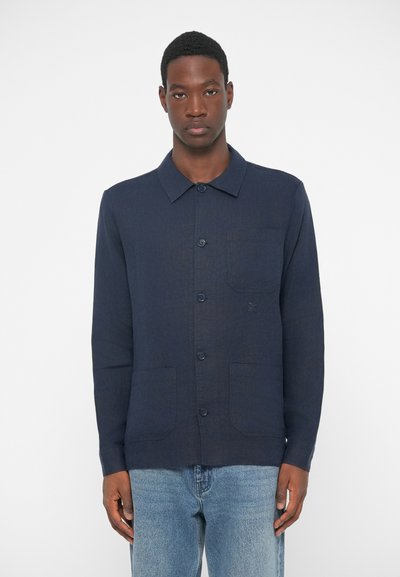 Mann in einem dunkelblauen Hemd mit Knöpfen und drei vorderen Taschen, trägt helle Jeans und steht vor einem schlichten weißen Hintergrund.