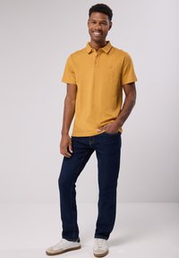 Polo shirt jaune moutarde à manches courtes avec trois boutons et un petit logo sur la poitrine, associé à un jean bleu foncé et des baskets blanches.