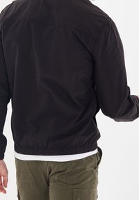 Veste noire en coton avec un design léger, taille élastique et détails comme des coutures et une chemise blanche contrastante en dessous.