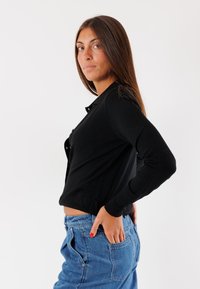 Cardigan nero a maniche lunghe con design aderente, caratterizzato da un collo rotondo e chiusura con bottoni. Abbinato a jeans blu a vita alta.