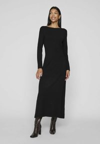 Abito maxi nero a maniche lunghe con scollatura a barca e silhouette dritta. Indossato con stivali lucidi fino al ginocchio, in piedi su uno sfondo grigio chiaro.