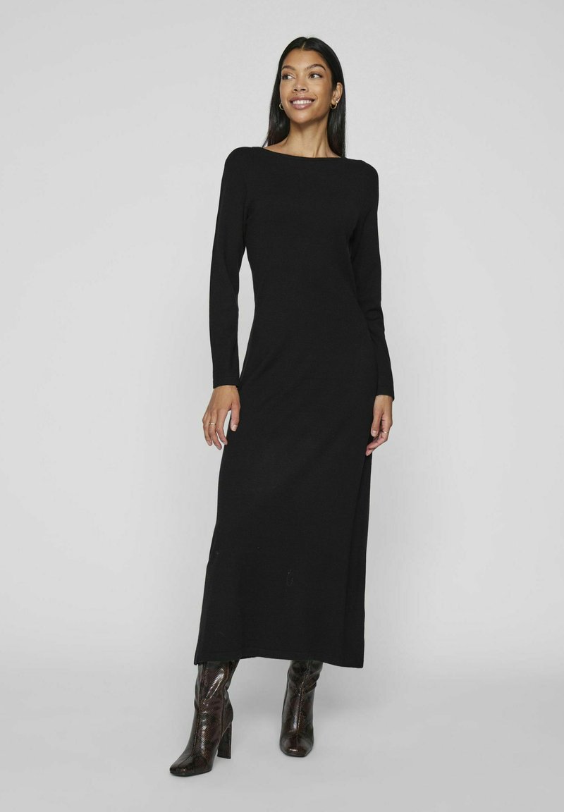 Abito maxi nero a maniche lunghe con scollatura a barca e silhouette dritta. Indossato con stivali lucidi fino al ginocchio, in piedi su uno sfondo grigio chiaro.
