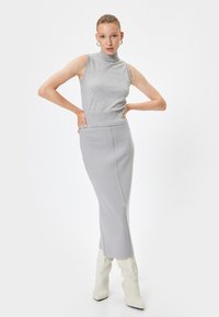 Grijze mouwloze coltrui gecombineerd met een lange grijze rok, met een strakke pasvorm en een zachte breistekstuur. Model draagt crèmekleurige laarzen.