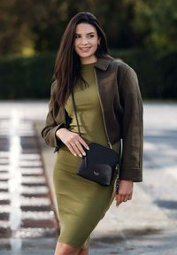 Černá kožená kabelka přes rameno se zlatým kováním. Olivově zelené žebrované šaty a olivově zelená bunda. Dlouhé tmavé vlasy, přirozené nehty a stříbrný prsten.