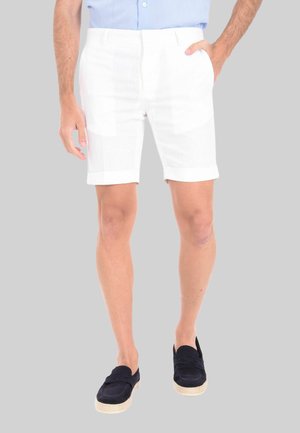 Uomo che indossa pantaloncini bianchi fino al ginocchio, camicia azzurro chiaro e scarpe slip-on nere, in piedi con una mano in tasca.
