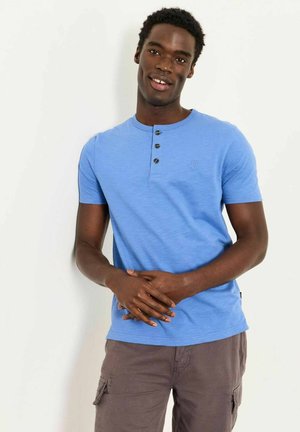 Mann steht vor weißer Wand, trägt blaues Henley-Shirt mit kurzen Ärmeln und graue Cargohose, Hände verschränkt, lächelt leicht.