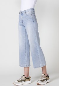 Vaqueros de pierna ancha de color azul claro con corte recto, hechos de denim, que cuentan con cinco bolsillos y un cierre de botón. Combinados con zapatillas blancas y doradas.