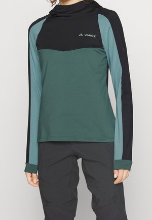 Osoba ubrana w zielono-czarną bluzę z kapturem z długim rękawem i czarne spodnie, pokazana od górnej części ciała do okolicy środkowej uda.