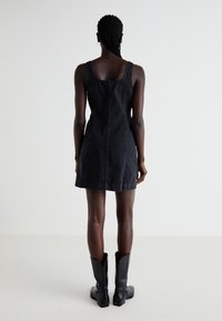 Robe en denim noir avec une coupe ajustée, décolleté carré et bretelles réglables. Présente des coutures visibles et une longueur au-dessus du genou. Bottes noires.