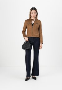 Veste cropped en daim marron avec poches zippées, assortie à un jean évasé noir et un sac à main noir. Des escarpins noirs pointus complètent le look.