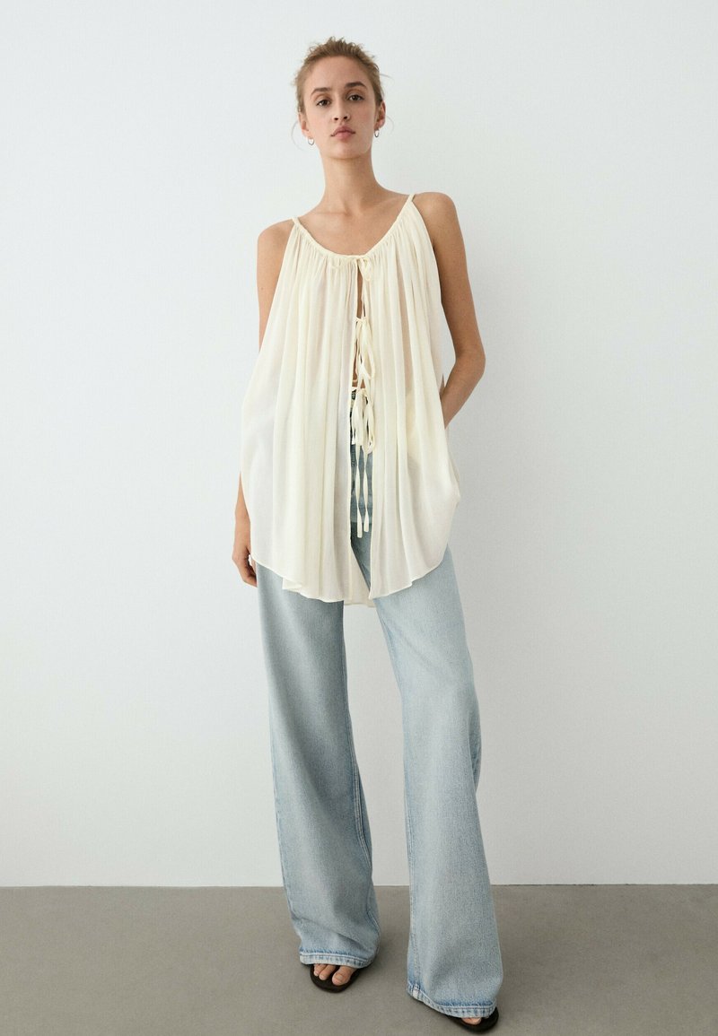 Vrouw die een crèmekleurige mouwloze geplooide blouse draagt met strikjes aan de voorkant en lichtblauwe wijde spijkerbroek staat tegen een effen witte muur.