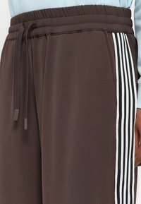 Braune Polyester-Sporthosen mit elastischem Bund und Kordelzug, die weiße gestreifte Akzente an den Seiten aufweisen.