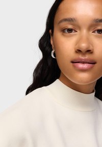 Boucles d'oreilles créoles rondes en argent texturé et pull à col roulé crème côtelé. Peau douce et lisse avec un maquillage naturel et des cheveux foncés ondulés.