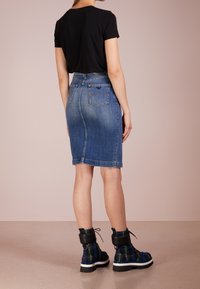 Žena v černém tričku, těsném modrém džínovém sukni ke kolenům a černých kostkovaných šněrovacích botách stojí na béžové podlaze.